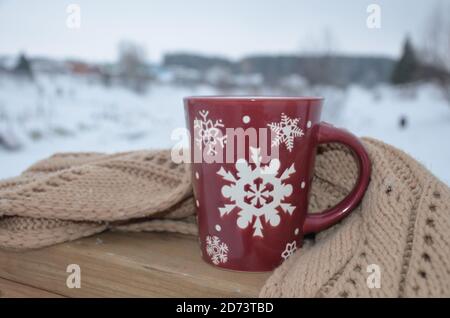 Coppa rossa o borgogna con un modello di fiocchi di neve sullo sfondo di una foresta invernale. Nevicare. Capodanno. Natale. Tazza con una bevanda calda al vento Foto Stock