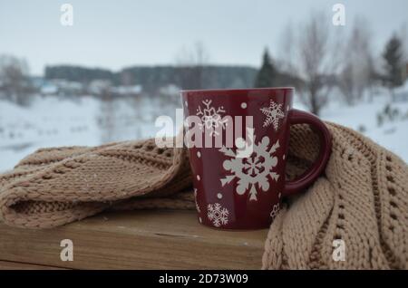 Coppa rossa o borgogna con un modello di fiocchi di neve sullo sfondo di una foresta invernale. Nevicare. Capodanno. Natale. Tazza con una bevanda calda al vento Foto Stock