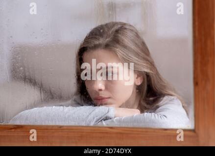 Infelice giovane donna che guarda fuori finestra con gocce di pioggia, sensazione di tristezza o malinconia Foto Stock