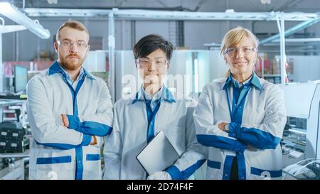 In High-Tech Factory: Ritratto di tre ingegneri professionisti che posano con le braccia incrociate. Nel gruppo scheda a circuito stampato elettronico di sfondo Foto Stock