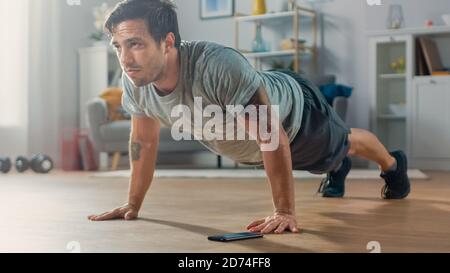 Atletico Fit Man in T-shirt e Shorts sta facendo esercizi Push Up mentre si usa un cronometro sul suo telefono. Sta addestrando a casa nella sua stanza vivente con Foto Stock