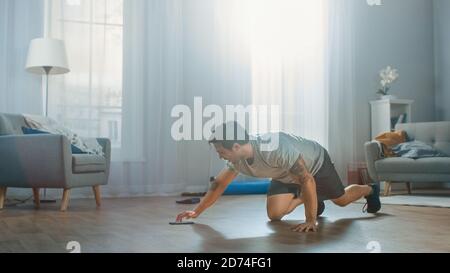 Atletico Fit Man in T-shirt e Shorts sta facendo gli esercizi alpinisti mentre si usa un cronometro sul suo telefono. Si sta allenando a casa nella sua brillante Foto Stock