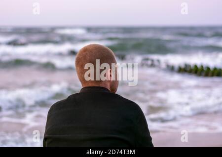 Il giovane si siede con la schiena in riva al mare al tramonto di sera. Messa a fuoco selettiva, sfocatura. Foto Stock