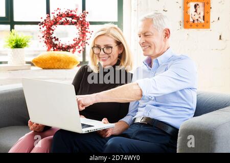 Foto di felice coppia senior utilizzando il computer portatile mentre si siede sul divano a casa e si naviga su Internet insieme. Uomo anziano che indica qualcosa sulla la Foto Stock