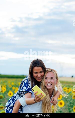 Un paio di giovani donne attraenti una bionda e il altre brunette che posano nei loro abiti firmati in un campo di girasoli utilizzando il loro smartphone Foto Stock