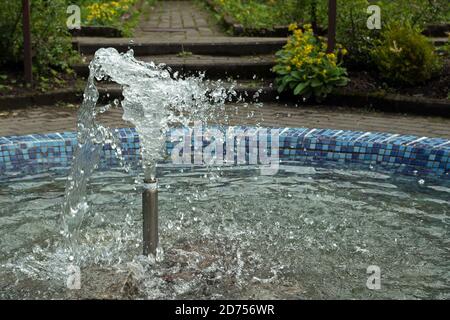 Fontana che spruzza acqua da ugello cilindrico in piscina circolare foderato con tessere mosaico in parco sullo sfondo di cespugli verdi e le fasi degli strati Foto Stock