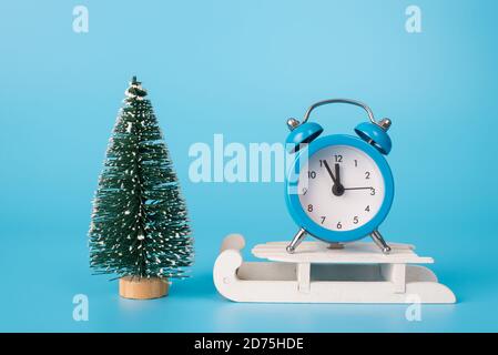 Cinque minuti prima del concetto di nuovo anno. Foto di orologio piccolo in piedi su slitte di legno bianco vicino giocattolo in miniatura abete sempreverde isolato sulla luce Foto Stock