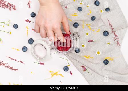 Sparato dall'alto di lamponi all'interno di un vaso con una mano di donna che ne raccoglie alcuni, su sfondo rosa con mirtilli, alcuni fiori selvatici e un panno Foto Stock