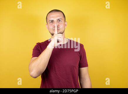 Uomo russo che indossa una t-shirt di base rossa su sfondo giallo insolato chiedendo di essere tranquillo con il dito sulle labbra. Silenzio e concetto segreto. Foto Stock