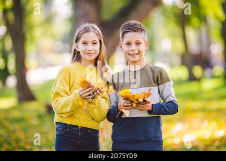 Felici gemelli adolescenti ragazzo e ragazza che si abbracciano l'un l'altro nel parco autunnale tenendo in mano foglie gialle cadute in tempo di sole. Tema della stagione autunnale Foto Stock