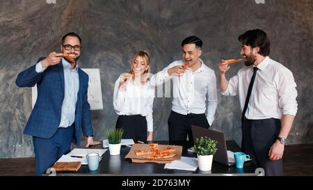 Felici i diversi colleghi trascorrono il lavoro pausa insieme mangiare take away consegna pizza in ufficio, sorridendo dipendenti multirazziali divertirsi, ridere e chattare Foto Stock