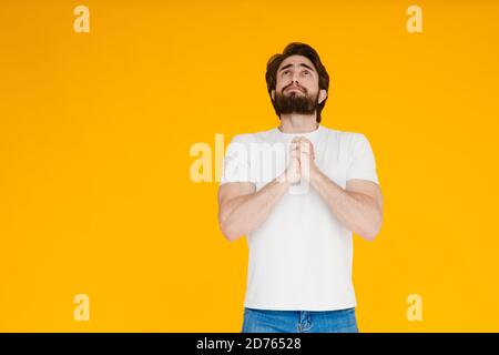 Ragazzo uomo con la bearded pleading in bianco t-shirt casual che posa isolato su parete gialla sfondo ritratto studio. Concetto di stile di vita di emozioni della gente Foto Stock