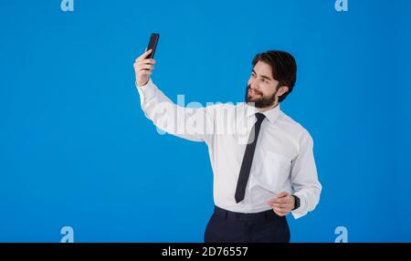 Un uomo d'affari giovane che parla al telefono su facetime . Un uomo con camicia bianca e cravatta scatta una foto selfie su sfondo blu Foto Stock