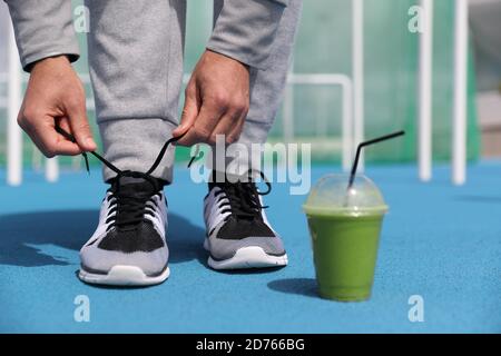 Palestra fitness man sempre pronto legando scarpe da running con frullato verde disintossicazione bevanda. Primo piano di allenatori da corsa con una tazza di plastica per la colazione Foto Stock
