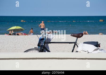 Una bicicletta lasciata appoggiata a un muro sul lungomare di Marotta, sotto il sole estivo. Alcuni turisti e il mare adriatico sullo sfondo Foto Stock