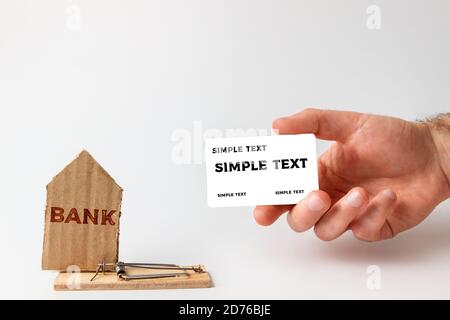 Concetto di investimenti e rischi bancari. La mano di un uomo tiene una carta di banca, e accanto ad essa è un mousetrap con una casa di cartone con una BANCA di testo. Simulazione, Foto Stock