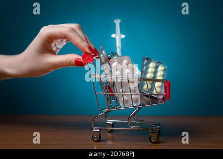 Una donna con una manicure rossa sta spingendo un piccolo carrello pieno di pillole e farmaci. Sfondo turchese. Primo piano. Concetto di prodotti farmaceutici, arredo Foto Stock