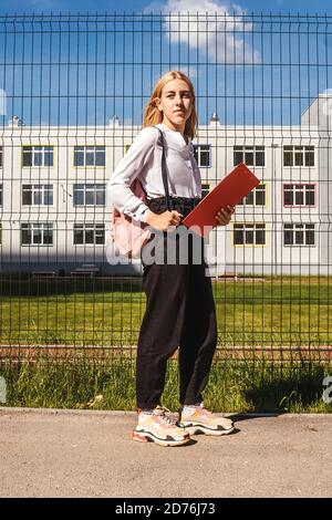 Giovane ragazza con cartella rossa vicino recinzione scuola. Ragazza in blusa bianca, zaino rosa. Foto Stock