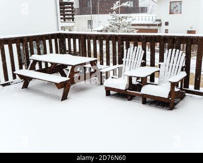 Due sedie in legno e un tavolo con panche si trovano su una veranda coperta di neve sotto una nevicata Foto Stock