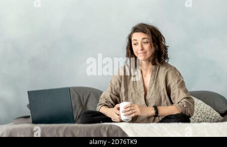Una giovane donna bruna usa un computer portatile nella sua camera da letto mentre si siede sul divano. Foto Stock