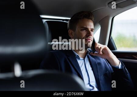 Primo piano ritratto di un giovane uomo in tuta che parla al telefono mentre si siede sul sedile posteriore dell'auto. Bel giovane uomo d'affari prova auto nuova Foto Stock