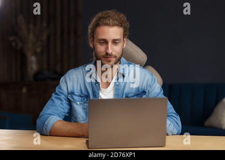 Ritratto di sorridente bel bell'uomo curly dai capelli lunghi che usa un laptop per una riunione online in videochiamata. Lavorare da casa. Comunicazione online con Foto Stock