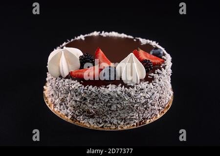 Vista in primo piano della gustosa torta marrone decorata con rasatura al cocco sui lati, bacche fresche e panna bianca. Splendido dessert delizioso con guarnizione al cioccolato isolata su sfondo nero. Foto Stock