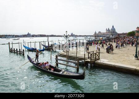 Turisti in gondola esplorando il canale veneziano, Venezia, Italia. Die Riva degli Schiavoni con stazione di servizio Gondola. Basilica di Santa Maria della Salute Foto Stock