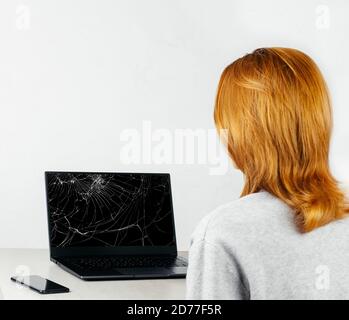 ragazza dai capelli rossi seduta a un tavolo e guardando un computer portatile con uno schermo rotto e rotto Foto Stock
