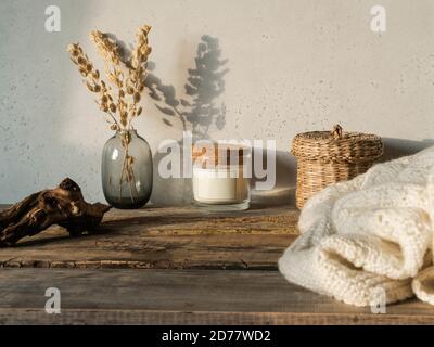 Accogliente casa d'autunno ancora vita di fiori secchi in vaso, cannuccia, candela di vetro, coperta a maglia su rustico tavolo di legno e ombre sulla parete dalla b Foto Stock