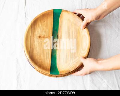 Mani femminili che tengono vassoi di artigianato rotondi in legno con inserto in resina verde su fondo tessile bianco. Vista dall'alto. Spazio di copia Foto Stock