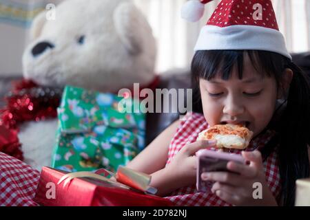 Felice bambina asiatica che mangia pizza e cerca uno smartphone in camera decorata per il nuovo anno. Cena di Natale fast food Foto Stock