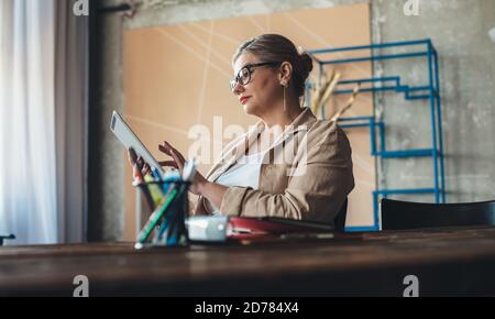 La donna caucasica anziana con gli occhiali sta usando una compressa durante una giornata di lavoro impegnativa da casa Foto Stock