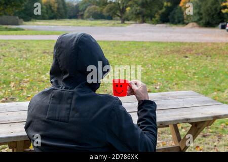 Uomo in cappotto nero con cappuccio, prendendo pausa bevendo una tazza di caffè da una tazza di plastica rossa con un sorriso su di esso. Seduto su una panchina ad un tavolo da picnic Foto Stock