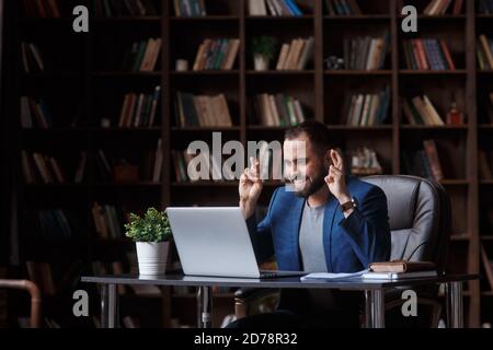 Un giovane uomo d'affari con la bearded nell'ufficio della biblioteca in un vestito blu guarda lo schermo del laptop e le attraversa le dita. Concetto di attesa per Foto Stock