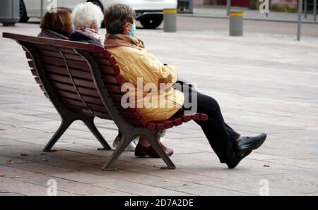 Tre Signore anziane con maschere di fronte seduta su una panchina nella piazza della città godendo pomeriggio all'esterno conforme con coronavirus e covid-19. Foto Stock