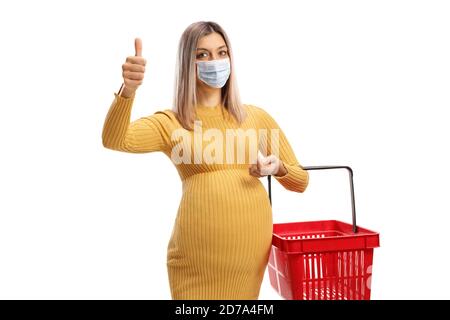 Donna incinta che tiene un cestino di shopping che mostra i pollici su e. indossare una maschera protettiva isolata su sfondo bianco Foto Stock