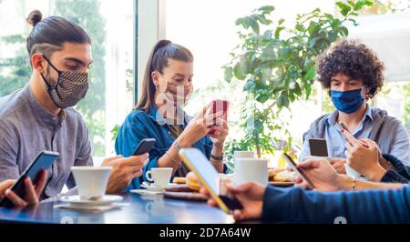un gruppo di amici sorridenti a colazione in un bar mentre interagiscono con la tecnologia. i compagni bevono caffè e mangiano mentre usano gli smartphone. Foto Stock