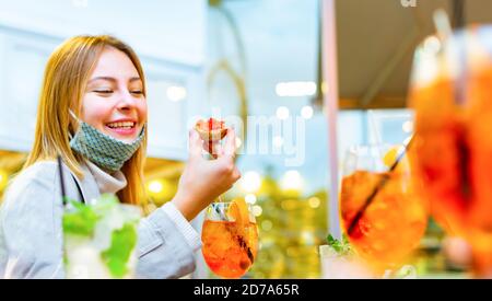 donna caucasica angelica sorride e prende un morso durante l'happy hour circondato da bevande colorate. ragazza bionda felice indossando maschera in un bar Foto Stock