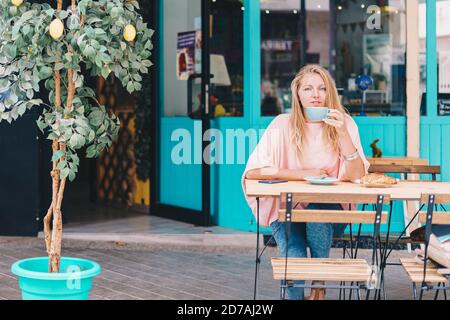 Bella donna bionda che guarda la macchina fotografica che fa colazione sulla terrazza di un caffè mentre beve il caffè. Foto Stock