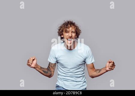 Stile libero. Uomo con capelli ricci in piedi isolati su mani grigie da parte in pugni eccitato Foto Stock