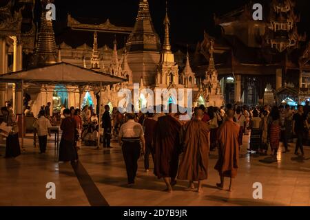 Yangon, Myanmar - 30 dicembre 2019: Tre monaci e visitatori passeggiate intorno alla pagoda Shwedagon e le sue strutture dorate circostanti di notte Foto Stock