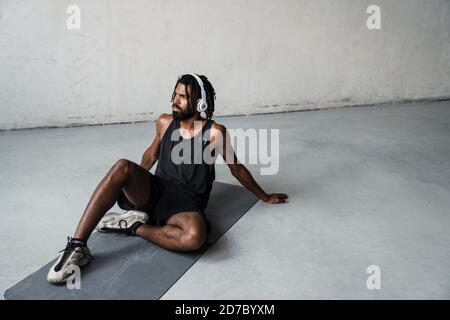 Immagine di un forte sportivo afroamericano che usa le cuffie mentre si è seduti su tappetino all'interno Foto Stock