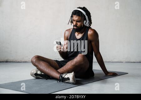 Immagine di sportivo afro-americano focalizzato utilizzando cuffie e cellulare mentre si siede sul tappetino all'interno Foto Stock