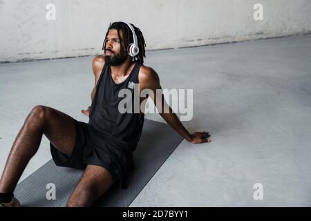 Immagine di un forte sportivo afroamericano che usa le cuffie mentre si è seduti su tappetino all'interno Foto Stock