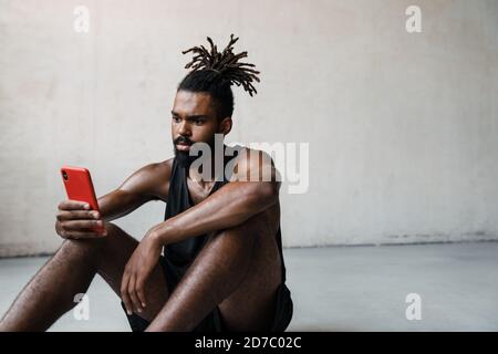 Immagine di giovane sportivo afroamericano che usa il telefono cellulare mentre posto a sedere al piano interno Foto Stock