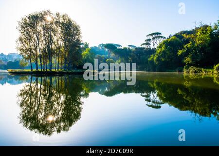 Gli alberi e il sole nel parco di Villa Ada a Roma si riflettono sulle acque del lago circostante creando un pattugliatore simmetrico. L'immagine presenta un leggero Foto Stock