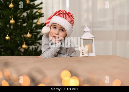 Un ragazzo con occhi tristi, seduto con la mano che gli tiene la guancia. Vestito in berretto. Mi sono appoggiato vicino all'albero di Natale. Foto Stock