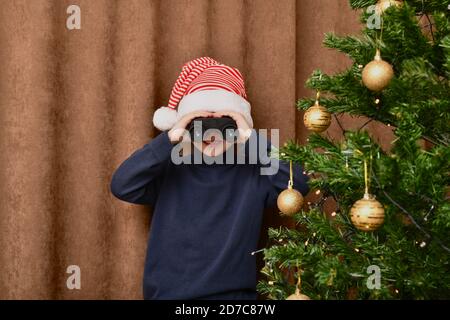 Il ragazzo sorride e guarda i regali vicino all'albero di Natale, guarda attraverso binocoli. Vestito con un carnivalcap. Foto Stock