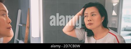 Donna matura asiatica di mezza età che tocca i suoi capelli guardando se stessa in bagno specchio panoramico banner stile di vita casa. Senior chinese signora capelli Foto Stock